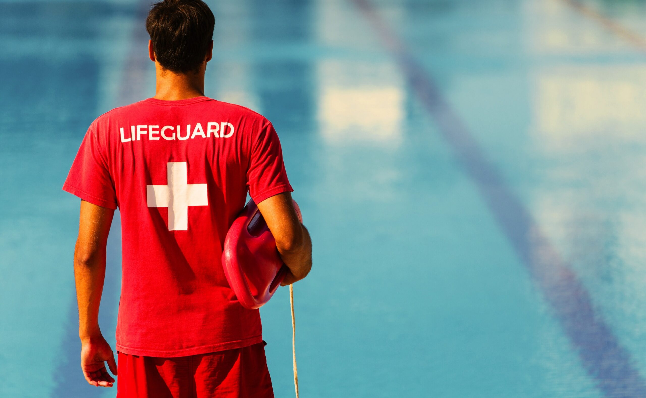 Rear view of male lifeguard in red uniform holding an emergency equipment watching the pool