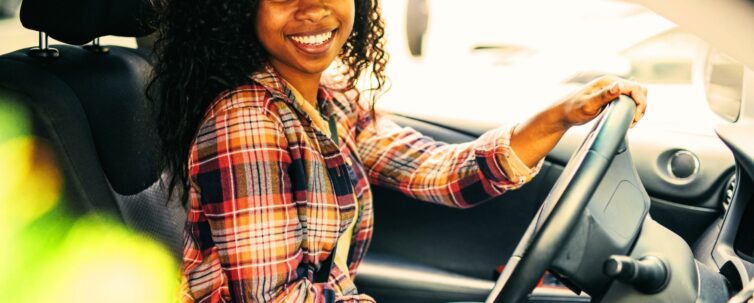 Millennial on driver side of car, holding the steering wheel smiling after the camera